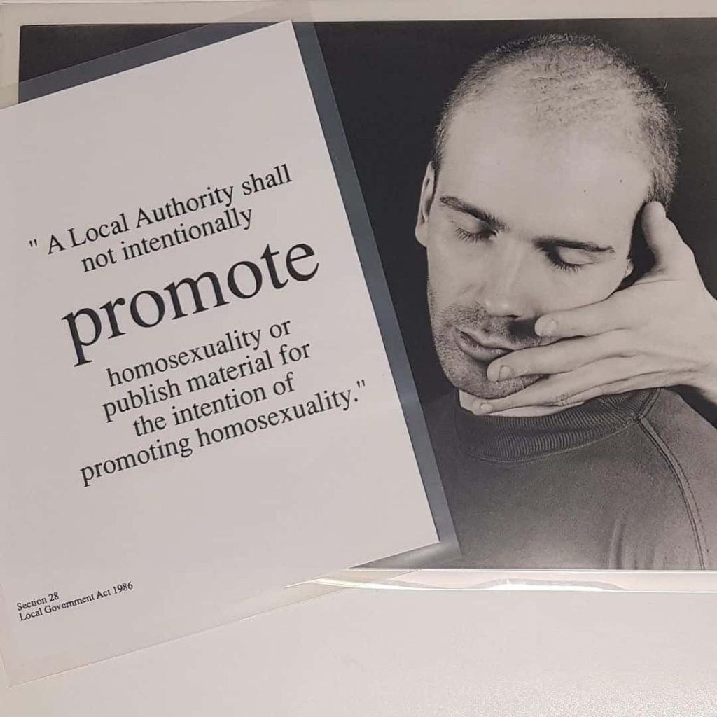 Black and grey photo of a photo with a laminated sheet of paper overlayed. The paper reads: "A Local Authority shall not intentionally promote homosexuality or publish material for the intention promoting homosexuality.". The photo show a peaceful but solemn young man leaning in to the caress of an open hand in the dark.