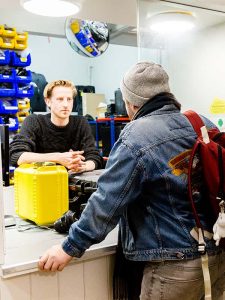 A student in denim jacket and grey beanie picks up equipment from the media resource. A bright yellow box sits on the counter and in the background there are racks of hardware and accessories for students to borrow.