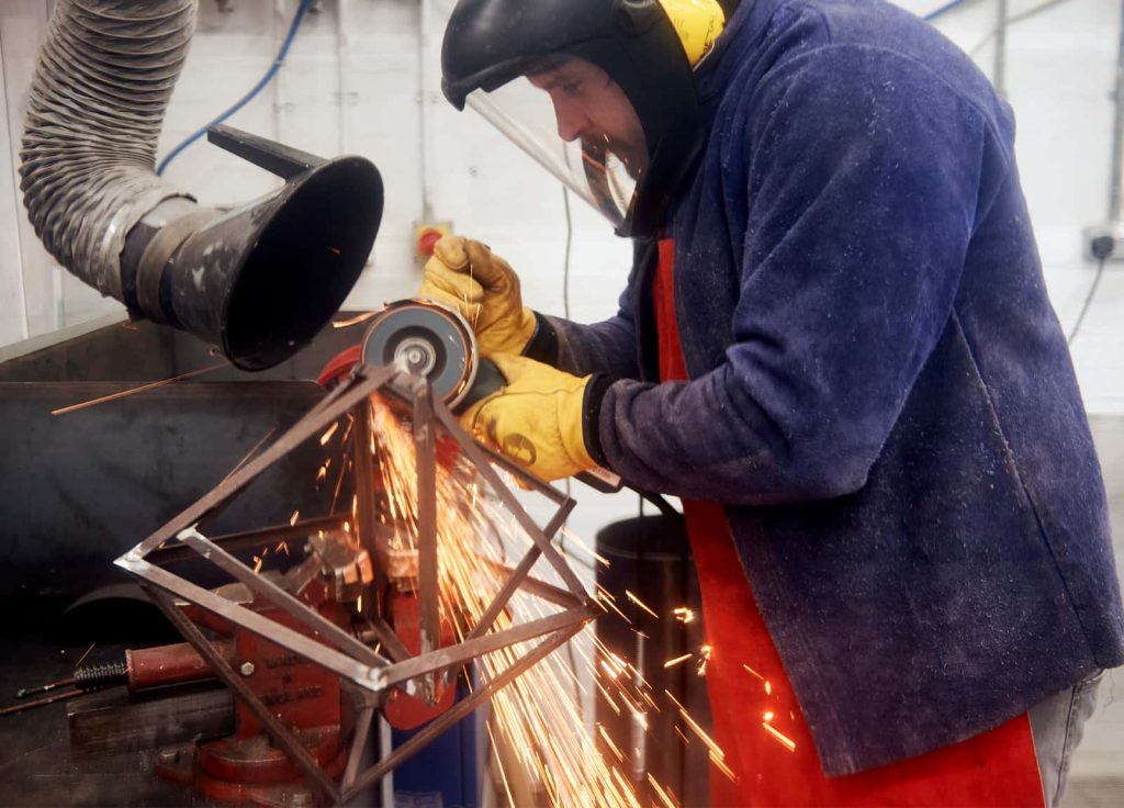 An Norwich University of the Arts Fine Art student with full-face visor and leather protective gear grinds down welds on a geometric metal sculpture in the 3D workshop, creating bright orange sparks