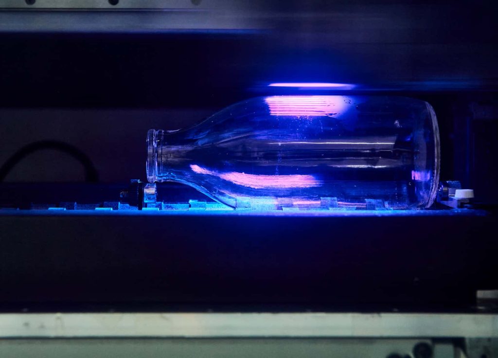 A clear glass bottle reflects vibrant violet light while being printed upon in the UV printer at Norwich University of the Arts