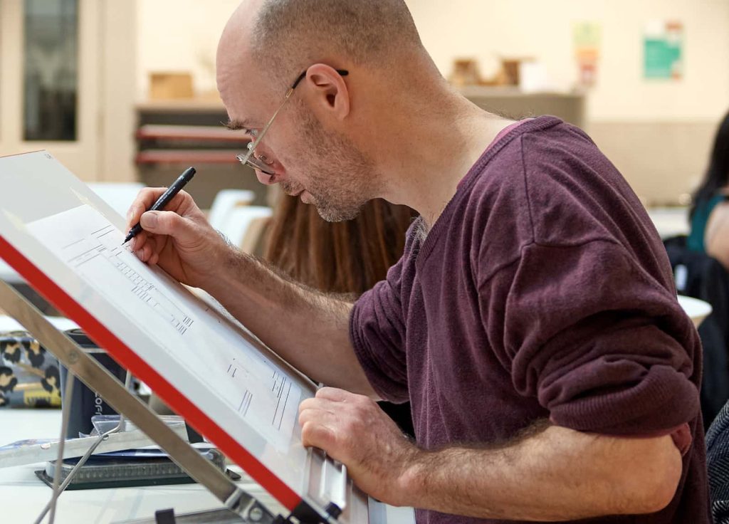 A mature student studying architecture at Norwich University of the Arts works intently on a technical drawing on one of the drawing tables in the architecture and interior design studio space.