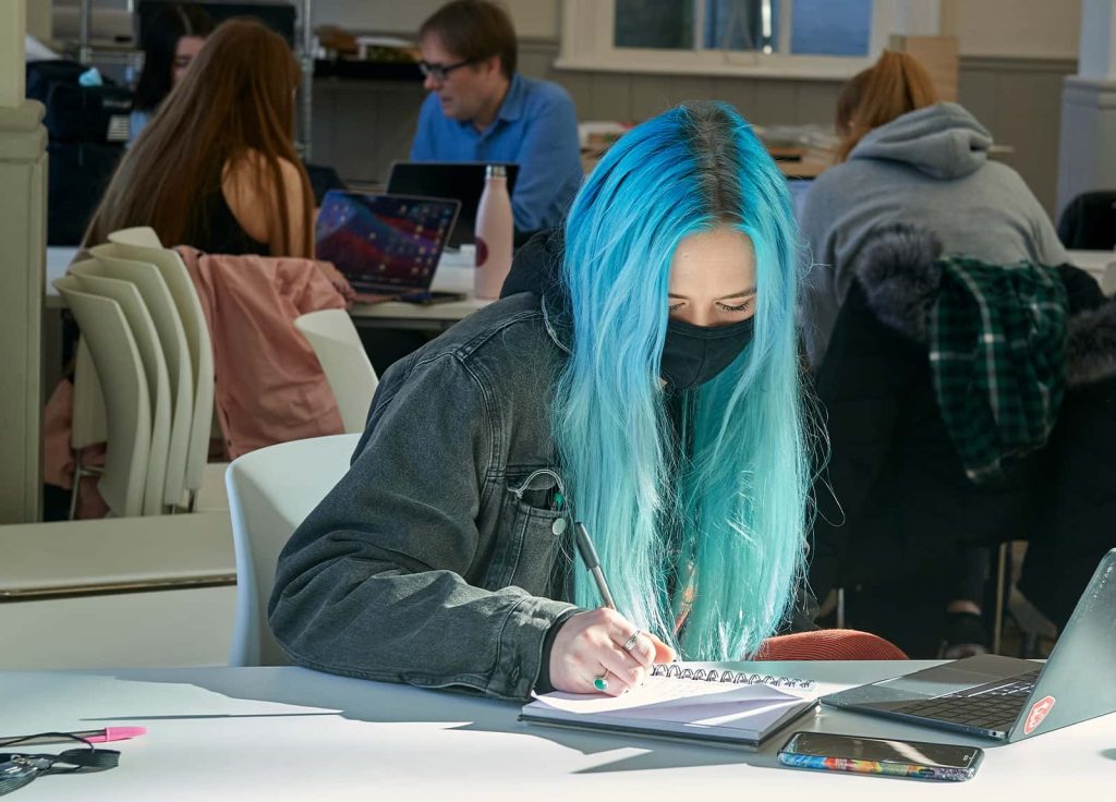 A student's long bright blue hair glows in a narrow beam of sunlight, while they take notes in the architecture and interior design studio at Norwich University of the Arts.