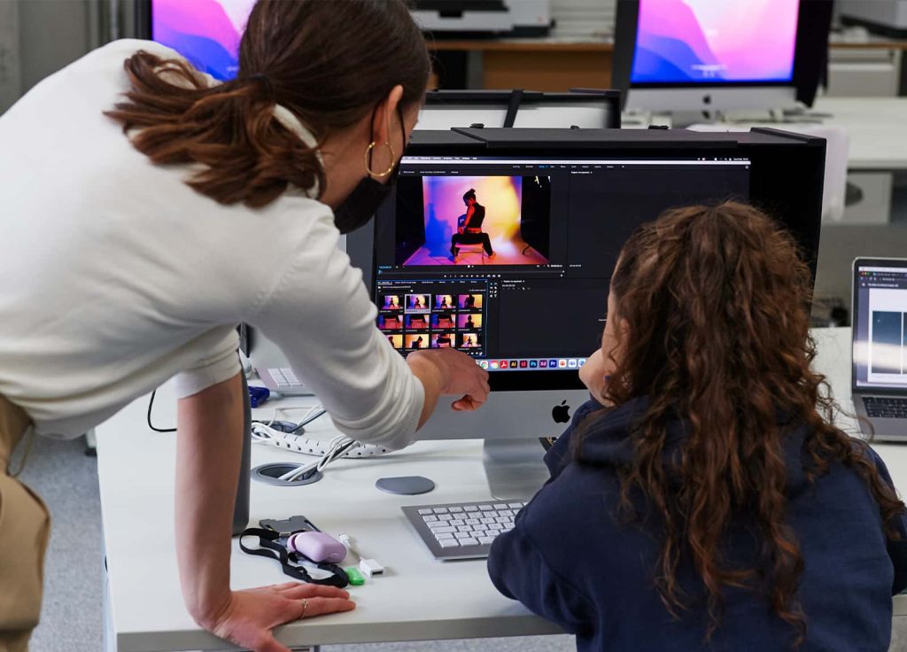 A member of staff gives 1 to 1 support to a student in the Digital Darkroom, who works on their images of a model sat facing away from the camera, with vibrant purple and yellow lighting. The student is working on a large iMac screen which has a black hood to protect the colour integrity seen.