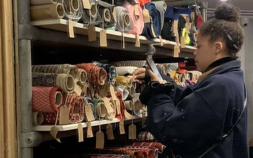 Photograph of BA Fashion student Zoe Brooker standing in front of shelving, stacked with rolls of fabric. Zoe is taking a photo of the fabric with her phone.