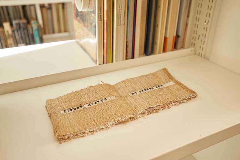 Work pictured resting on shelf. Work is in the shape of a folded book made with brown fabric that reads: flower, drives my green age