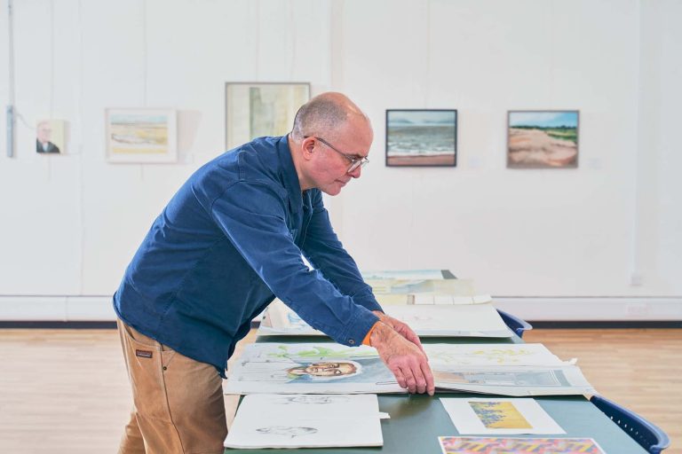 Will Jefferies in a blue shirt and brown trousers looking at portfolios of his own work next to a row of framed paintings on a wall