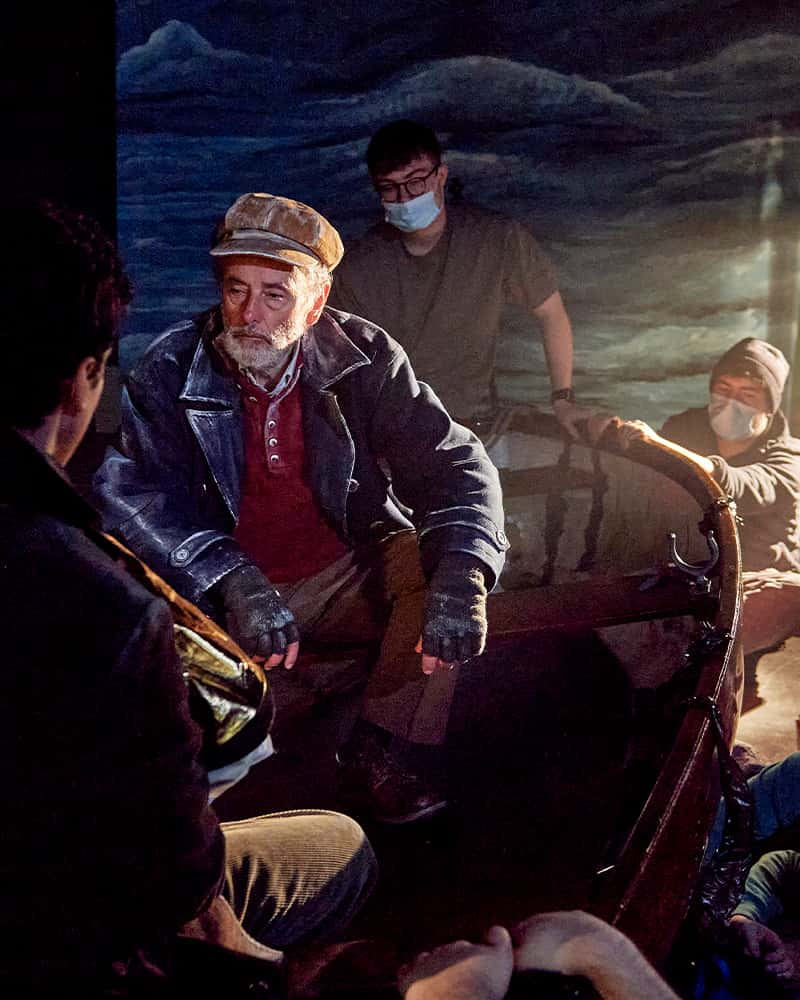 An old sailor sits in a rowing boat with a short grey beard. The boat is being hand rocked by students who crouch out of shot, in front of a large painted backdrop of stormy skies and seas. The sailors short peaked hat, jacket, and fingerless gloves have been drybrushed with white paint around the edges and details which makes him look slightly like he is a painting, and a beam of harsh warm light obscures some details in the photo.