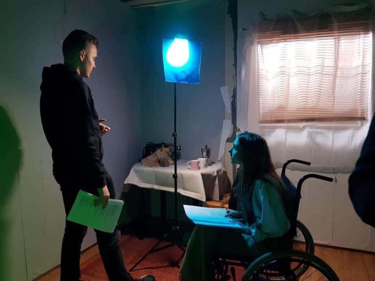 Image of two people reading scripts in a dark room in a house in a film set. The curtains are closed and there is a table with refreashments on. One person is in a wheelchair.