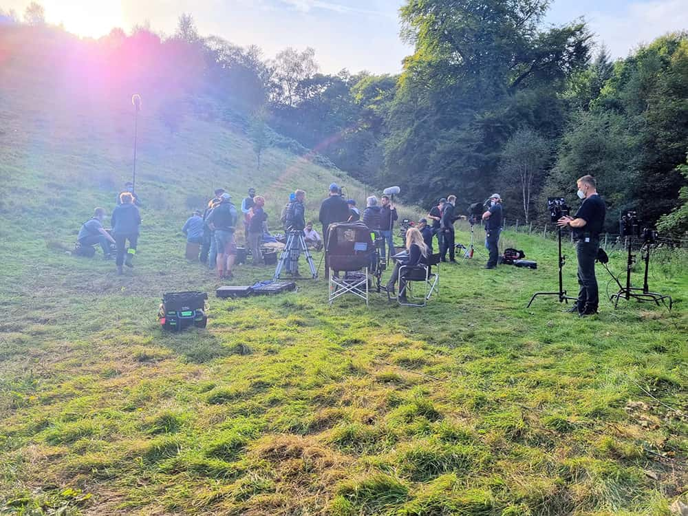 Image showing a group of cast member and crew on a film set in a field with a forest in the background.