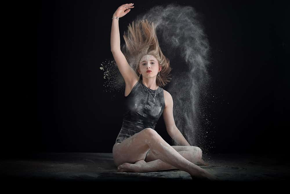 Image showing person sitting crossed legs in a dark room wearing a black sleeveless leotard. The person is throwing white powder over their head and their hair has been tossed upwards.