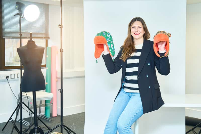 BA Fashion Marketing and Business lecturer and owner of Little Hotdog Watson, Emma Thompson, is pictured perched on the end of an empty white table, inside a fashion studio. Emma is holding both hands up, in each hand is a vibrant coloured children's hat from her own brand