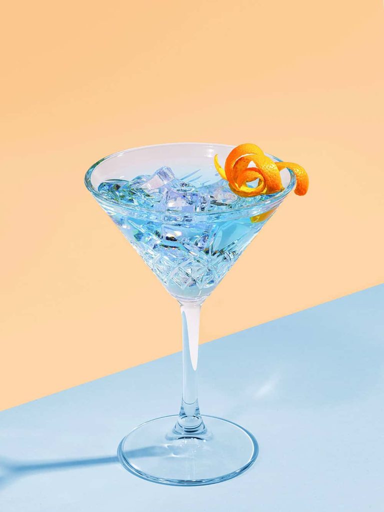 Photograph showing martini glass on a blue table with ice and blue liquid with an orange peel on the rim. Image is shot against an orange backdrop.