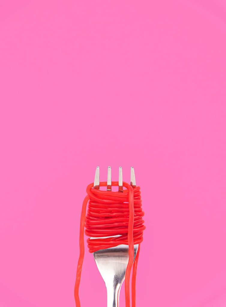 A silver fork on a bright pink background. The fork has a red strawberry lace wound around it like spaghetti