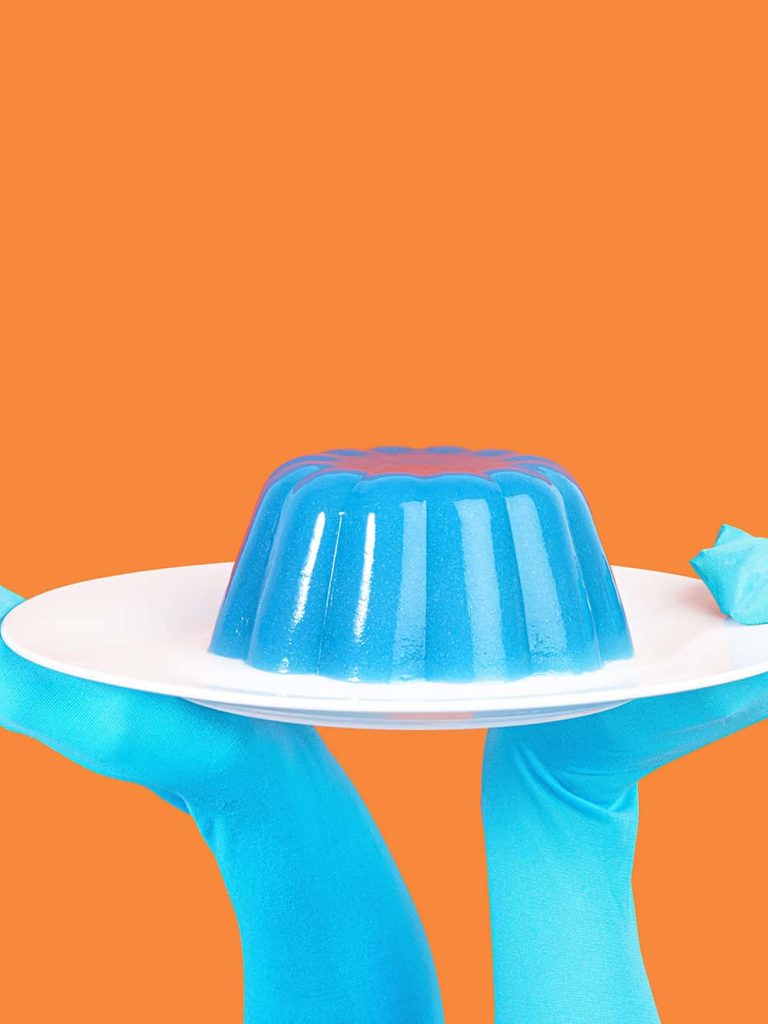 Photograph showing a person with blue hands holding a white plate with blue jelly. Shot on an orange backdrop.