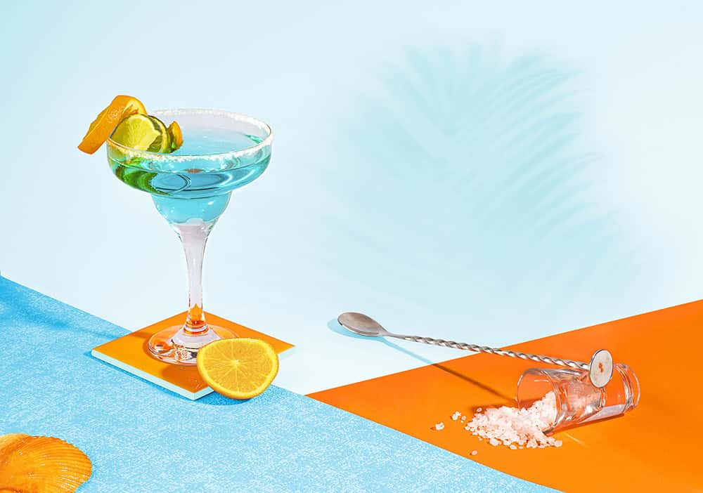 Photograph showing martini glass on a blue table with ice and blue liquid with an orange peel on the rim. Image is shot against an orange and blue backdrop.