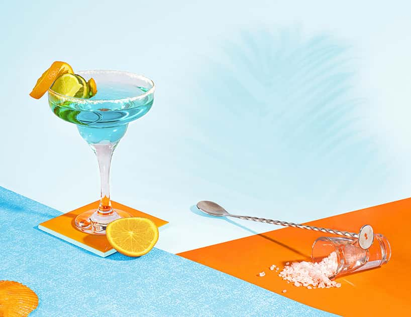 Photograph showing martini glass on a blue table with ice and blue liquid with an orange peel on the rim. Image is shot against an orange and blue backdrop.