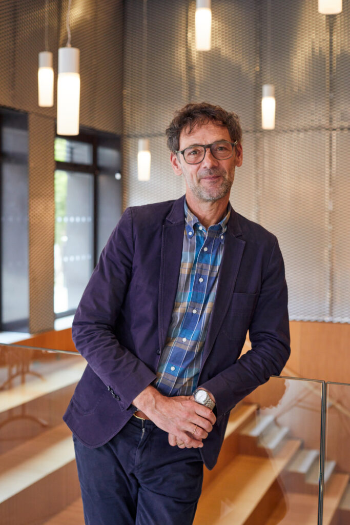 Professor Louis Nixon leaning against a glass partition, wearing a navy jacket and trousers with a colourful check shirt.