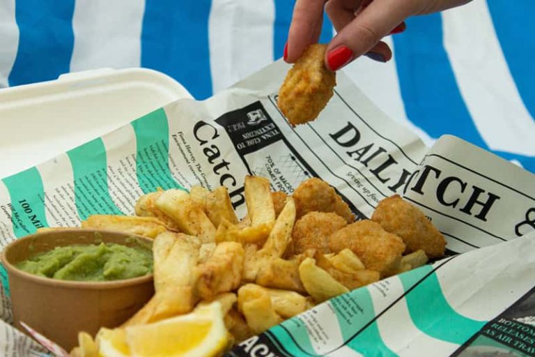 A photograph of vegan breaded scampi and chips, in traditional newspaper, with a cardboard tub of mushy peas. A hand is picking up a piece of vegan scampi. The newspaper is printed with articles on overfishing, to raise awareness of the damage caused to our oceans.