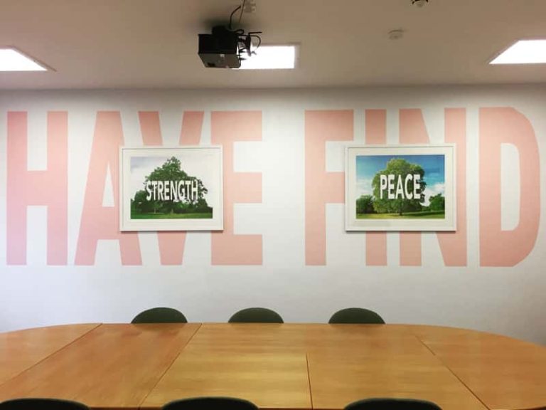 Photograph of a conference room with a large table and chairs in the centre. On the walls is large lettering, spelling out 'Have' and 'Find', coloured pale pink. In two white picture frame centred over each of the words, is a photograph of a tree, with the word 'Strength' in one frame, and the other the word 'Peace'. The work was created by Carl Rowe, for Norwich University of the Arts' research output.