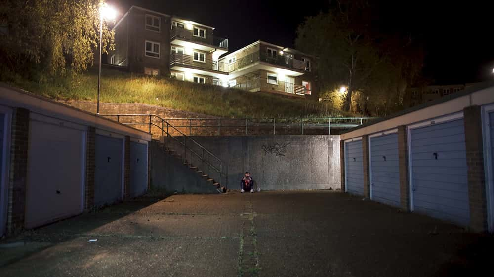 Image showing person sitting against a wall on a dimly lit street with car garages