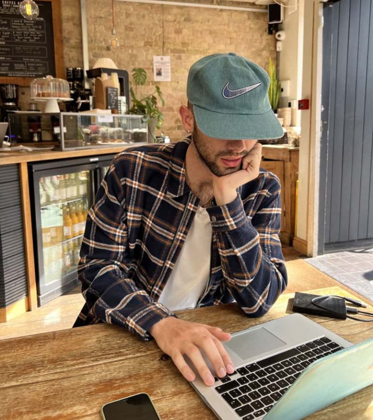 BA Fashion Communication and Promotion student Joe Corlito is sat at a wooden table in a modern cafe, working on his laptop. He is looking down at his screen, wearing a sage green baseball cap that covers half of his face