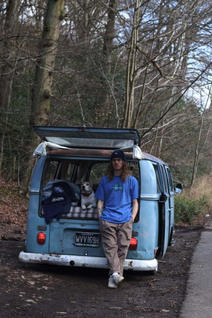 A photograph of a sky blue imported Volkswagen Type 2 camper van, parked in front of trees at the side of a road, with the boot open. Inside the van is a small mattress, with a white American Bulldog laying on top. Outside the car, owner Jack is leaning against the back, next to the dog. He is wearing a blue Stussy t-shirt, with camel coloured chinos.