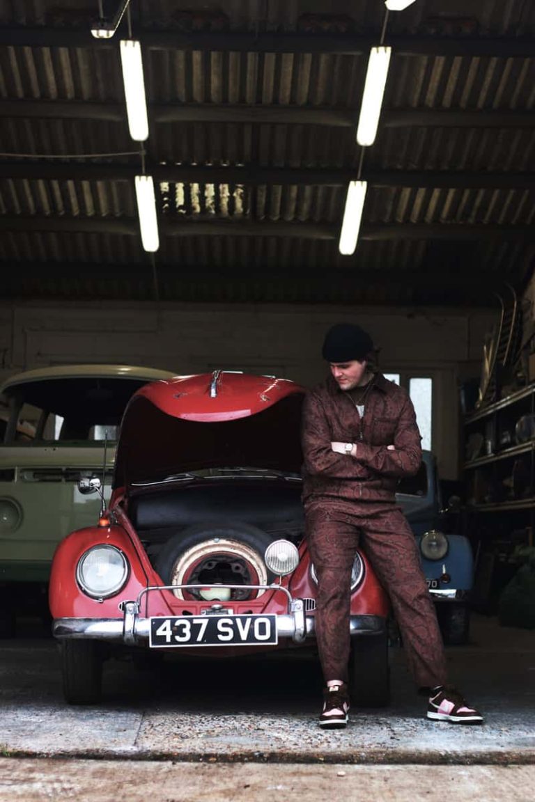 A photograph of a classic red Volkswagen beetle parked inside a warehouse garage, with the bonnet popped open. Leaning against the car is a male model wearing a maroon paisley patterned Stussy tracksuit, with a black beanie hat. He is looking sideways down towards his car