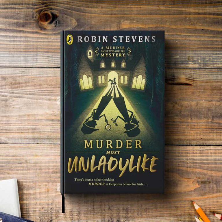 A top-down photograph of a hardback book on a wooden tabletop. The book cover illustrates a mysterious mansion in the distance, with two figures illuminated in the doorway. Their shadows are reflected down the path, where you can see them holding magnifying glasses and dressed as detectives. At the bottom of the book cover is the title; A Murder Most Unladylike