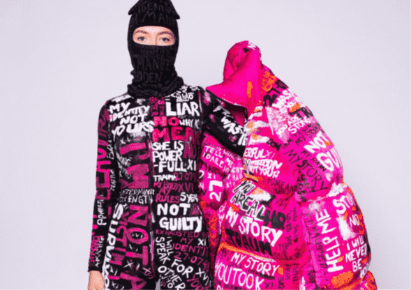 The model is swamped in an enormous ankle-length pink puffer jacket, which is covered in bold handwritten phrases and statements.. They are wearing a black balaclava, and lace-up boots.