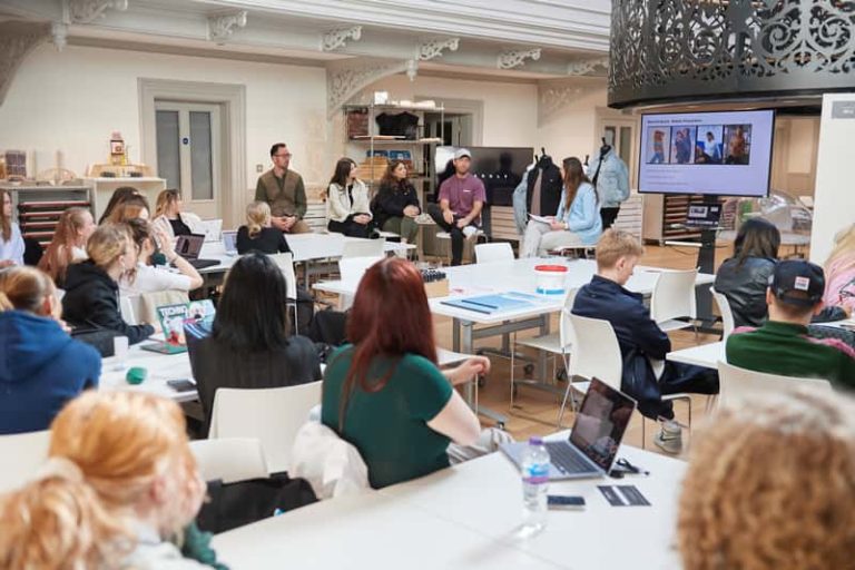 A photograph of the interior design studio at Boardman House, Norwich. A TV screen is placed near the staircase in the centre of the room, and has a presentation of fashion images from clothing brand Blakeley. Sat next to the TV are Norwich University of the Arts staff and Blakeley employees. They are presenting a project brief to a class of Interior Design and Fashion Marketing and Business students