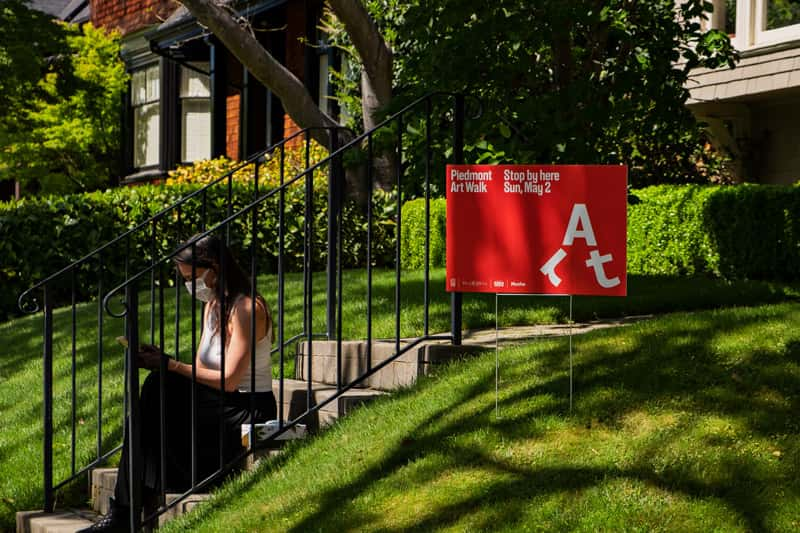 A photograph of a woman sitting on some steps outside a house. In the grass is a red sign advertising Piedmont Art Walk