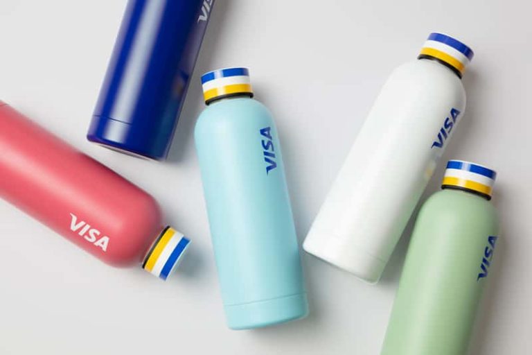 A photograph of five insulated water bottles, in a coral pink, navy blue, pale blue, white, and mint colour. The bottles are scattered on a grey background. Each bottle cap has a blue, white and yellow stripe. On the side of the bottle is the Visa logo.