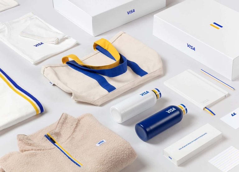 Flatlay of various merchandise, including tote bag, reusable bottle, a folded beige zip-up fleece, folded white t-shirt, pencil, notebook and cardboard packing boxes. All white, with a blue and yellow stripe, for the VISA rebrand