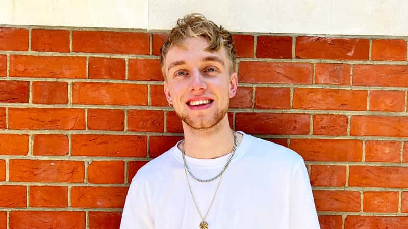 A photograph of BA Graphic Design alumni Tim Wilcox. Tim is standing in front of a red brick wall, wearing a white t-shirt, looking slightly past the camera