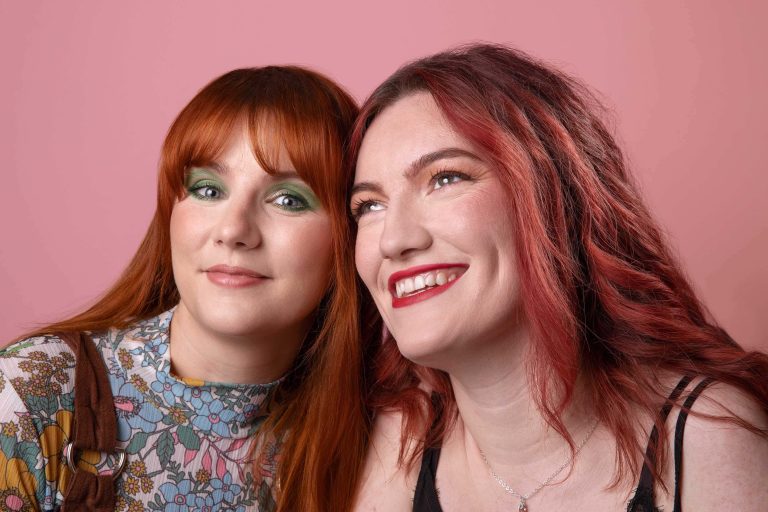 Photograph of two people with red and orange hair in front of a pink background starring at the camera