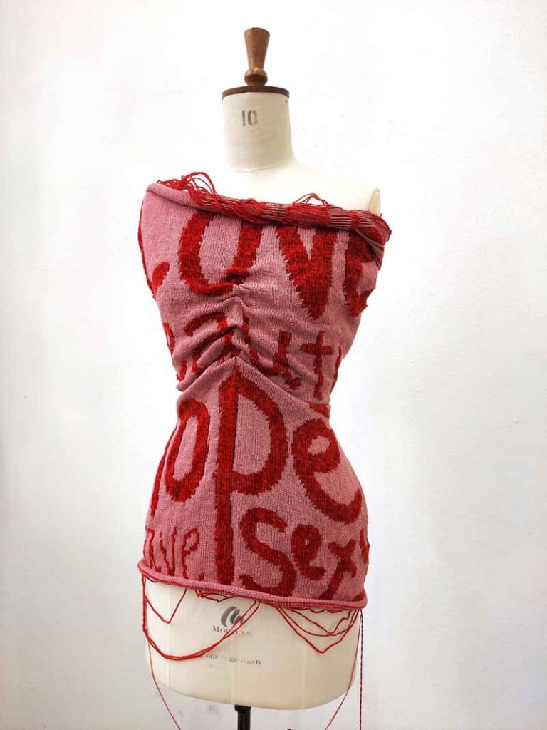 A fashion mannequin torso, with a pink knitted bandeau dress around the shoulders, town to the hips. In a red yarn, weaved into the dress are the words 'love', 'hope', 'sex'