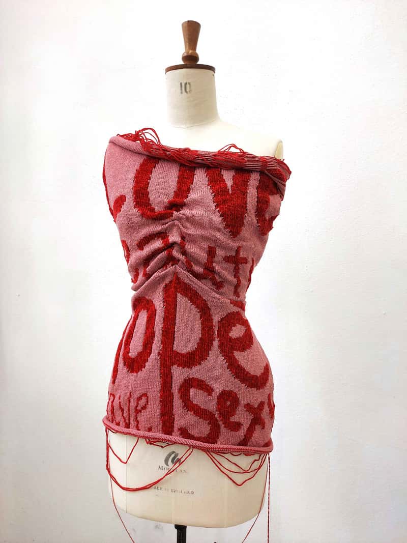 A fashion mannequin torso, with a pink knitted bandeau dress around the shoulders, town to the hips. In a red yarn, weaved into the dress are the words 'love', 'hope', 'sex'