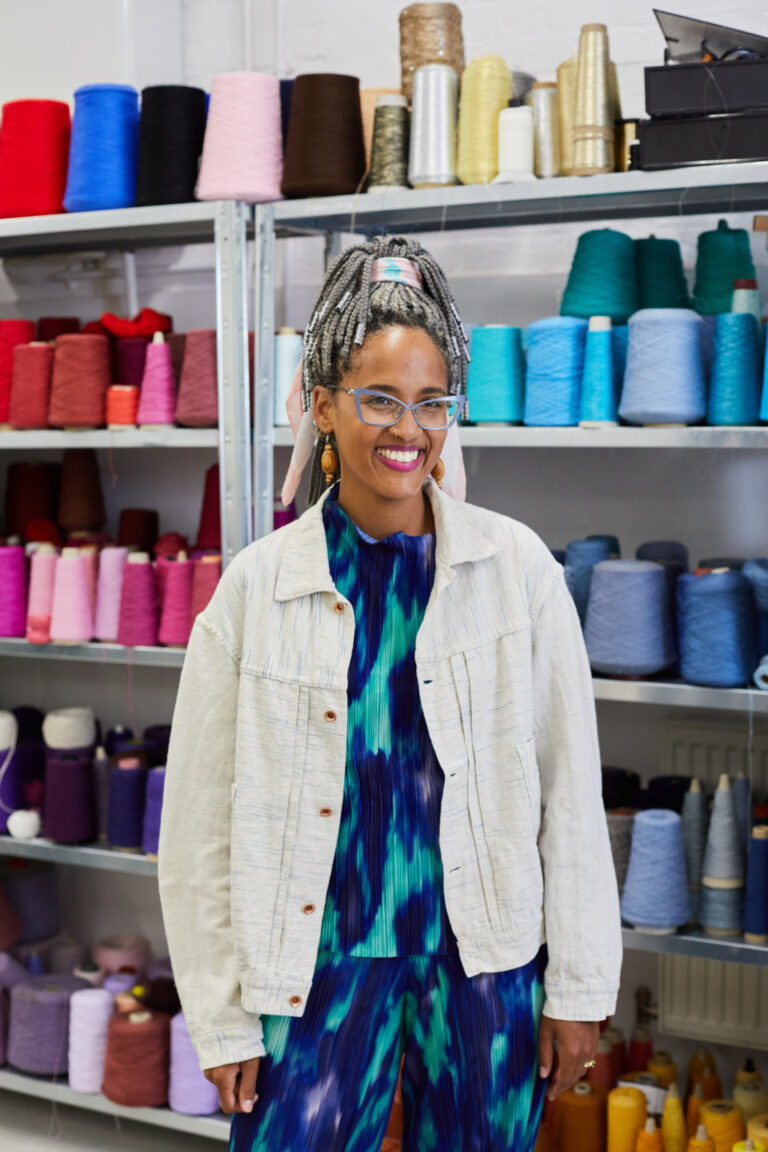 Senior Lecturer Odette Steele stands for a photo in front of shelves which are full of colourful reels of thread and yarn. Odette wears pastel cat-eye frame glasses, and a light blue denim jacket over dark blue tie-die pattern top