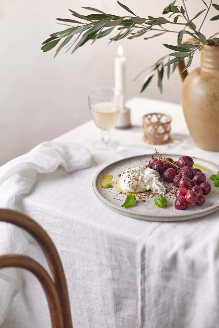 Photograph showing well laid dinner table with candle, plant and table cloth, with plat of burette and grapes.