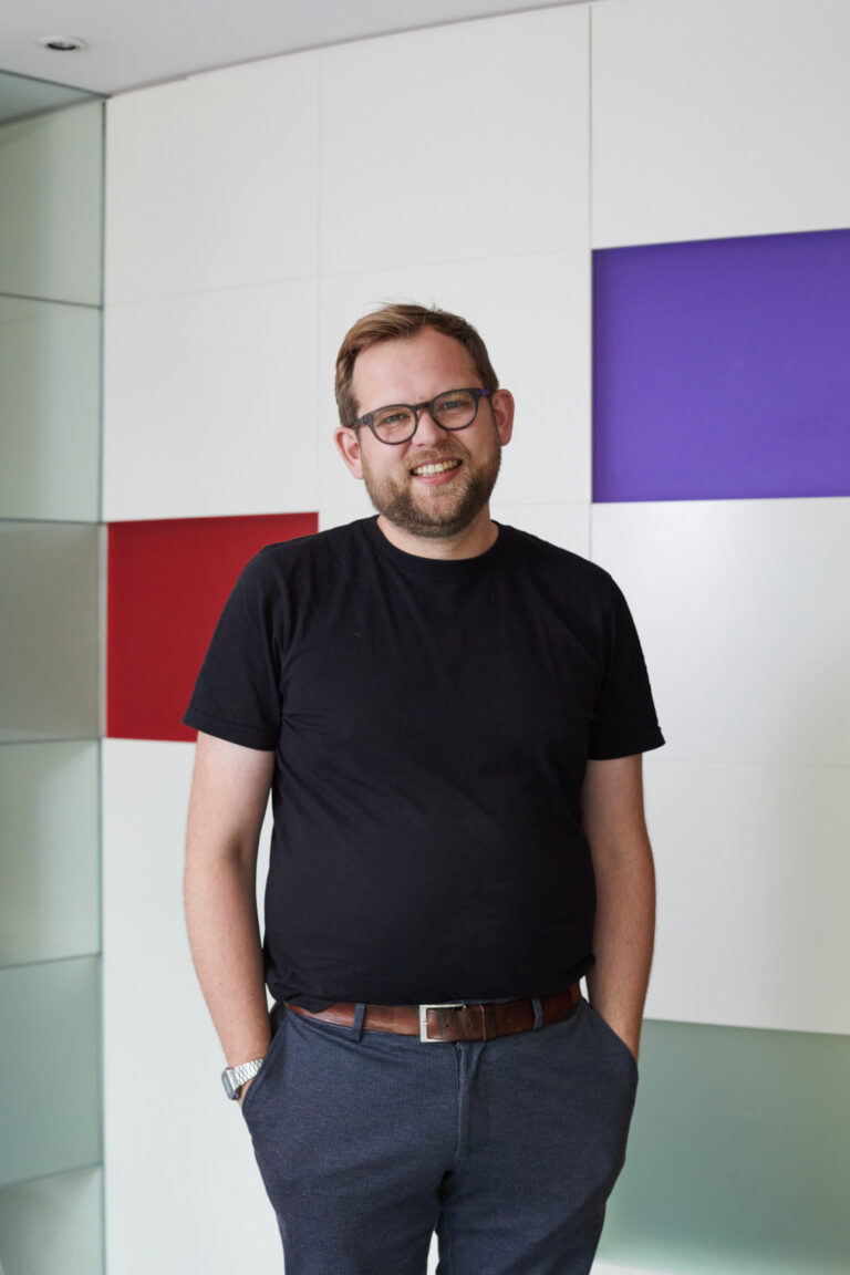 Portrait of Games Art and Design Lecturer and Industry Liaison Jake Montanarini . Jake wears a fitted black Tee tucked into smart navy trousers, and stands with his hands in his pockets.