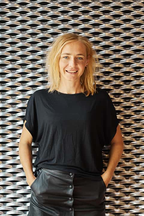 Acting Lecturer Dr Ilona Krawczyk staff portrait. Ilona stands in front of some decorative metal walls in the Duke Street Riverside building. Ilona has straight shoulder length blonde hair, and wears a casual fit black tee with short sleeves and a black leather skirt.