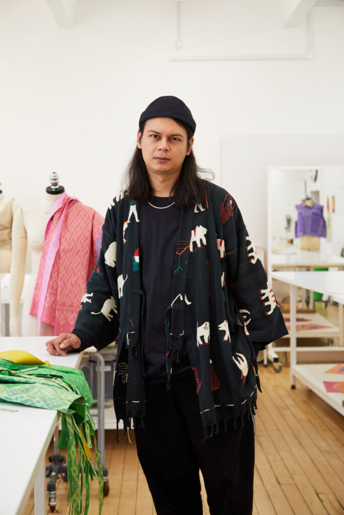 Fashion Course Leader River Chadprajong-Smith stands in a Fashion workshop with some colourful fabrics. River has shoulder length black hair, and wears a black beanie, loose black t-shirt, black trousers, a silver chain, and an open black cardigan patterned with ignorant style animal illustrations