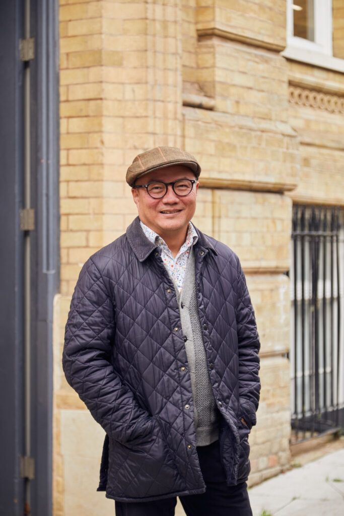 Architecture Lecturer William Chen stands in front of the St Georges building at Norwich University of the Arts. William wears a flat peaked cap and navy quilted jacket