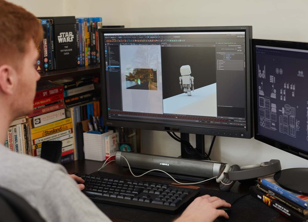 A student working with two screens in their accommodation, the desk is dark wood and has a bookshelf beside it, with books stored in stacks as well as vertical.