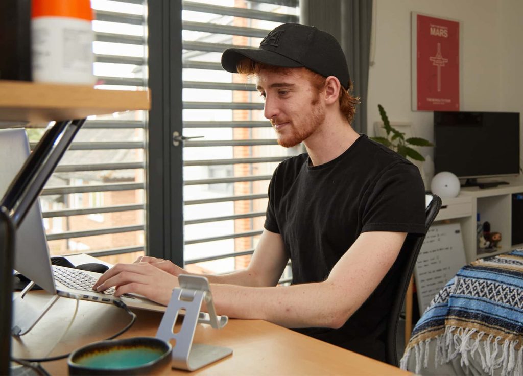 A student working at a laptop in their accommodation. The laptop is on an aluminium stand, in the background there is a plant and a TV on a bookshelf