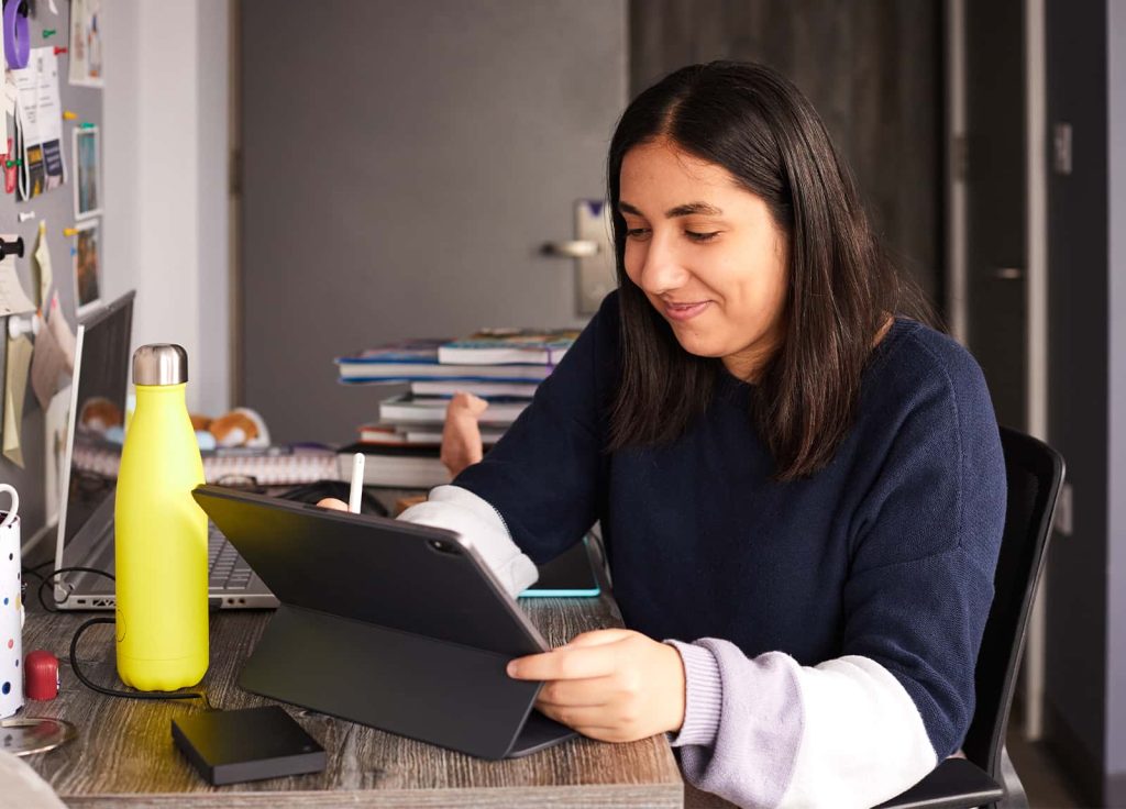 A student working with a stylus on a tablet device at a desk in their accommodation, the desk is a dark grey wood and also has a laptop and books on it