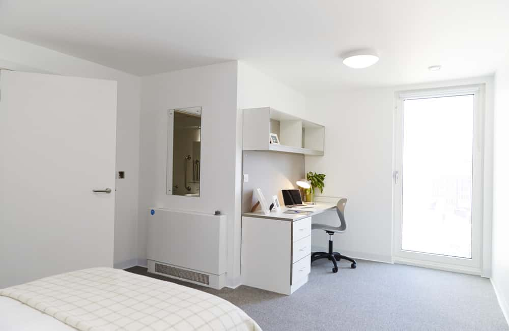 A large, modern bedroom in the All Saints Green accommodation at Norwich University of the Arts. The room has white walls, furniture and ceiling, and a light grey carpet