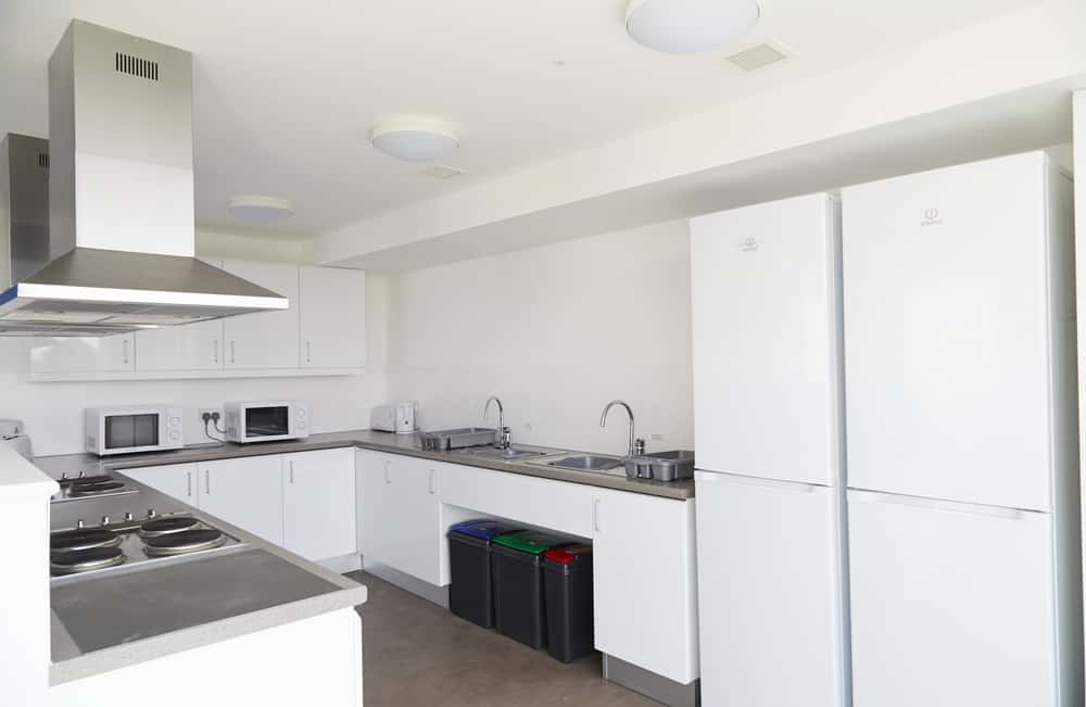 A large, modern kitchen in the All Saints Green accommodation at Norwich University of the Arts. The kitchen has two large fridge freezers, several microwaves and hob units, recycling bins, metal extractor hoods mounted to the ceiling. has white walls, furniture and ceiling, and a light grey carpet