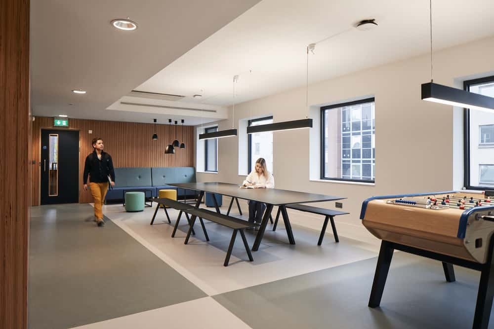 Students in a large open plan communal space with long tables, hanging lights, and football table in the Duke Street Riverside accommodation at Norwich University of the Arts