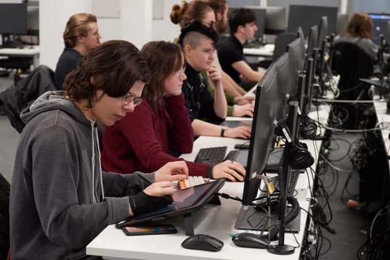 A photograph of a row of computers taken from the end of the table. Students are sat at each computer working individually on digital projects